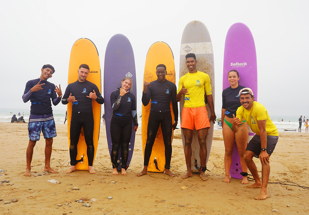 Group of guests at Nomad Surf Camp Morocco posing with surfboards in colorful lycra wetsuits on the beach